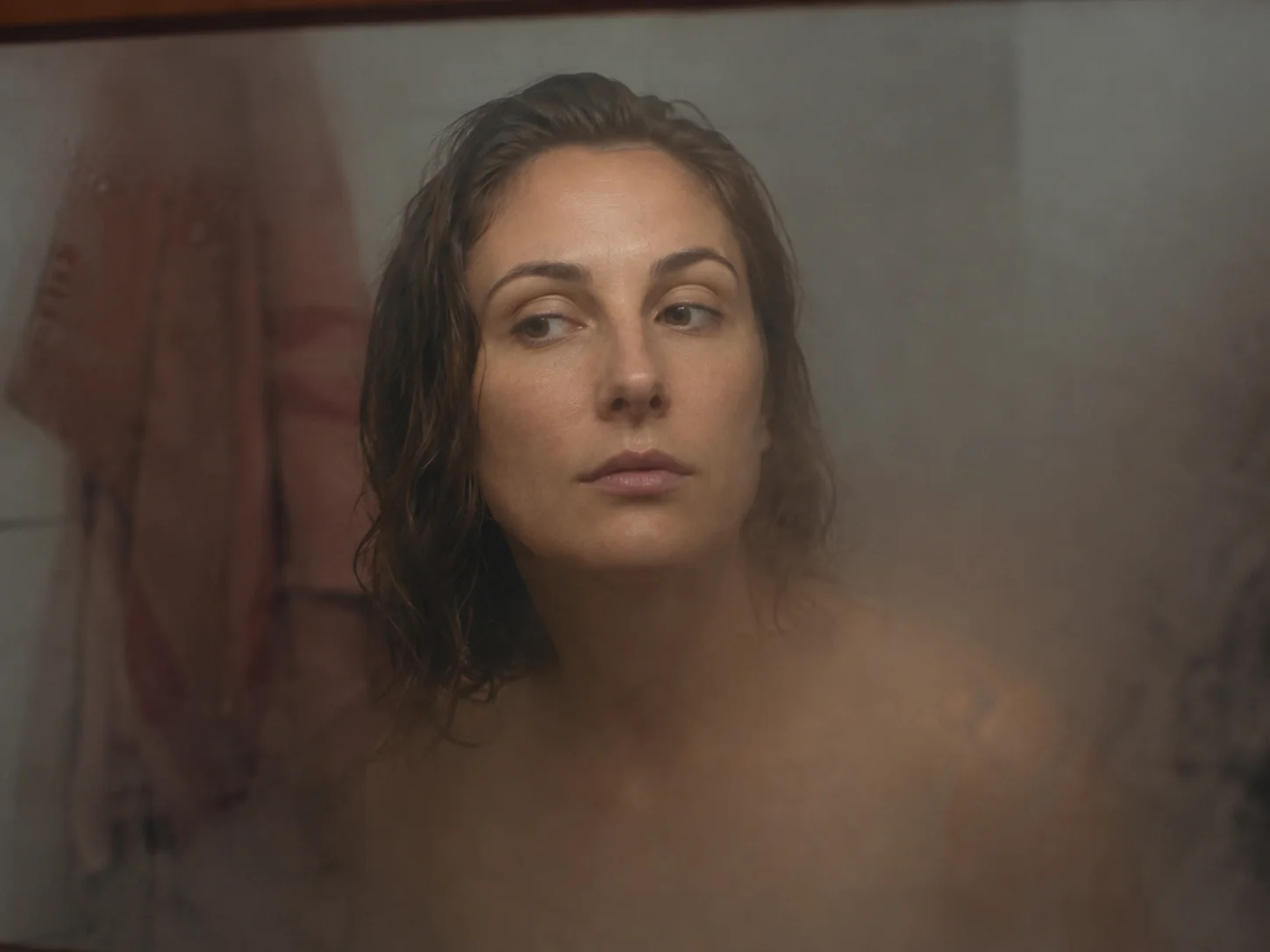 A woman glancing at her reflection in a dim, steamed-up bathroom mirror while getting ready.
