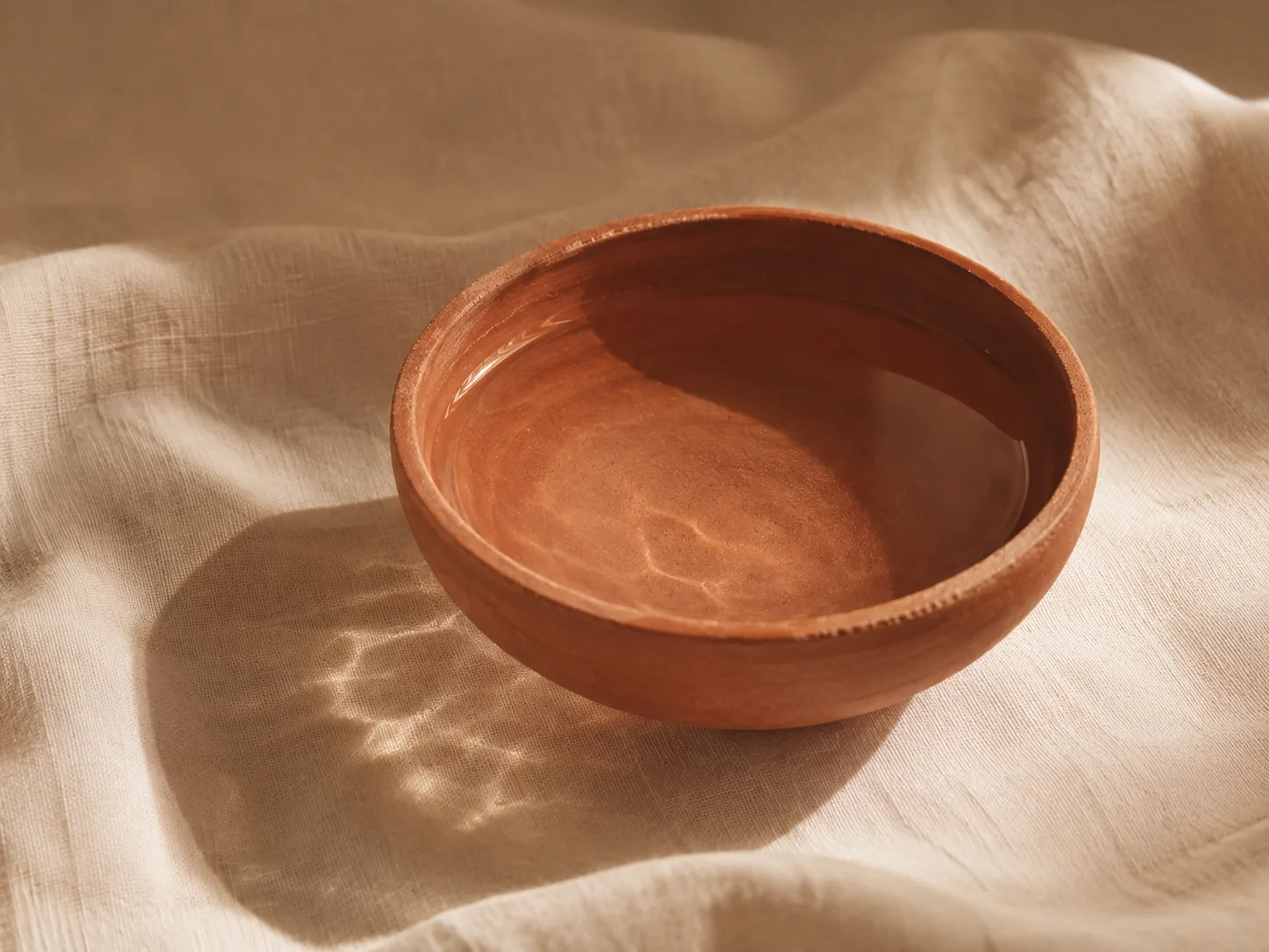 A shallow terracotta bowl of water on linen, the surface catching low afternoon light.